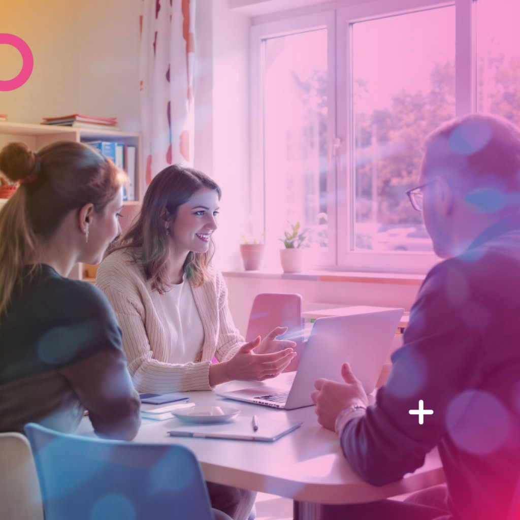 חדר מואר עם חלון גדול. שלושה אנשים יושבים סביב שולחן עגול עם מחשב נייד פתוח ומחברות. אישה מחייכת ומסבירה, ושני האחרים מקשיבים לה. התאורה ורודה־סגלגלה עם אפקטים דיגיטליים, היוצרים אווירה מודרנית וחדשנית